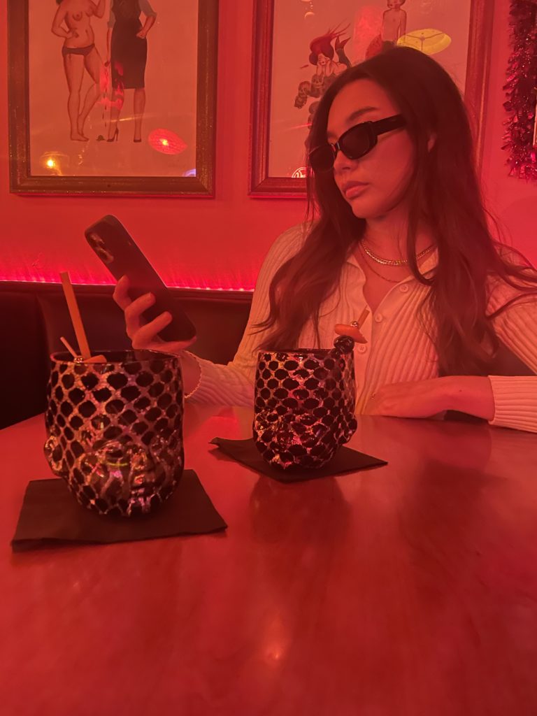 Dark red bar scene with a female modle checking her phone with a Baby Head Cup in the foreground.